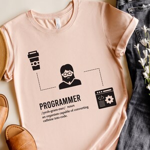 Könnte beinhalten: Ein pinkfarbenes T-Shirt mit einem schwarzen Grafikdesign, auf dem "PROGRAMMIERER /proh-gram-mer/ Substantiv ein Organismus, der in der Lage ist, Koffein in Code umzuwandeln" steht. Die Grafik enthält eine Kaffeetasse, eine Person mit Brille und Bart und einen Computerbildschirm mit Code.
