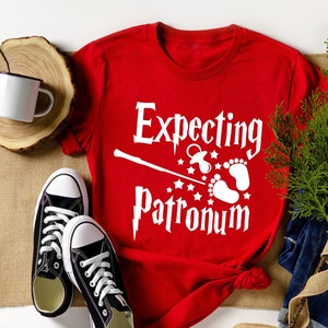 May include: Red t-shirt with white text that says "Expecting Patronum" with a wand, a pacifier, and baby footprints.