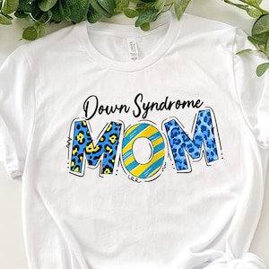 Op de afbeelding: Wit T-shirt met de tekst "Down Syndrome MOM" in blauwe, gele en zwarte gepatroneerde letters.
