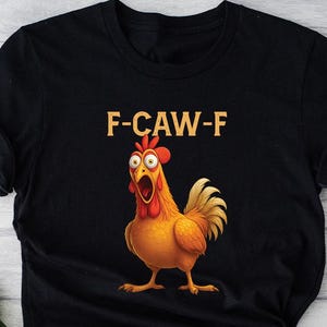 May include: Black t-shirt with a cartoon rooster graphic and the text "F-CAW-F" in yellow. The rooster is depicted with an open beak, orange and yellow feathers, and a red comb. The shirt has a round neck and short sleeves.