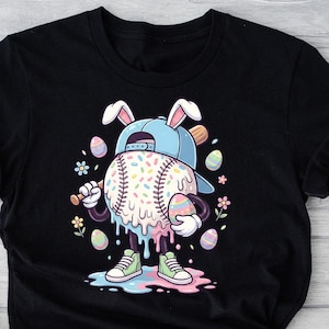 Puede incluir: Camiseta negra con un personaje de béisbol con orejas de conejo y gorra, sosteniendo un huevo de Pascua. La pelota de béisbol está decorada con chispas. El diseño incluye colores pastel y acentos florales. Una prenda casual.