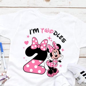 May include: White toddler t-shirt featuring Minnie Mouse with a pink polka dot bow and dress. The number 2 is in black with pink polka dots. Text reads "I'm TwoDles". White sneakers and colorful markers are also in the image.
