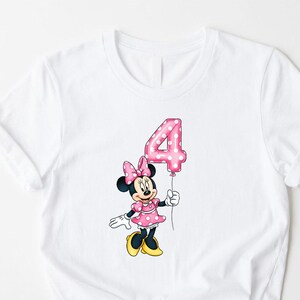 K&ouml;nnte beinhalten: Wei&szlig;es T-Shirt mit einer Cartoon-Minnie-Maus, die einen rosa Ballon mit der Zahl 4 h&auml;lt. Minnie Mouse tr&auml;gt ein rosa Kleid mit wei&szlig;en Punkten, eine passende Schleife und gelbe Schuhe. Das Design befindet sich mittig auf der Vorderseite des Shirts.