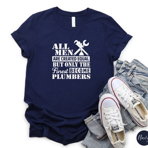 May include: Navy blue t-shirt with white text that reads "All men are created equal but only the finest become plumbers". The shirt features a white wrench and pipe graphic.