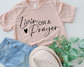 Peut inclure: Un t-shirt rose pâle avec le texte "Livin on a Prayer" en écriture noire avec un petit cœur à gauche du mot "Livin".