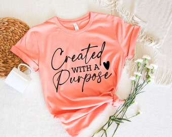 Puede incluir: Una camiseta de color rosa coral con el texto "Created with a Purpose" en letras negras y un pequeño corazón negro.