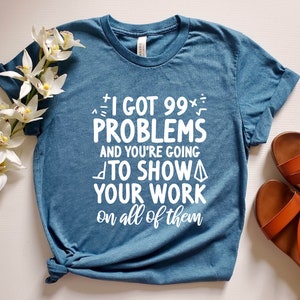 Op de afbeelding: Een blauw T-shirt met witte tekst die luidt "Ik heb 99 problemen en je gaat je werk op alle laten zien". De tekst is omringd door wiskundige symbolen.
