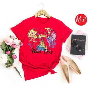 K&ouml;nnte beinhalten: Ein rotes T-Shirt mit einer farbenfrohen Grafik mit Micky-Maus-Ohren, einem Schloss und dem Text "Your Text". Das Design umfasst Farbspritzer in Gelb, Lila und Blau. Das Shirt wird auf einem Kleiderb&uuml;gel pr&auml;sentiert.