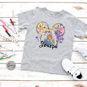 K&ouml;nnte beinhalten: Graues Kurzarm-T-Shirt mit einem farbenfrohen Aufdruck eines Schlosses und Mickey Mouse-Ohren mit dem Namen "Joseph" in schwarzer Schrift. Das Design enth&auml;lt Spritzer in Lila, Gelb und Rosa. Wei&szlig;e Turnschuhe und Marker sind ebenfalls sichtbar.