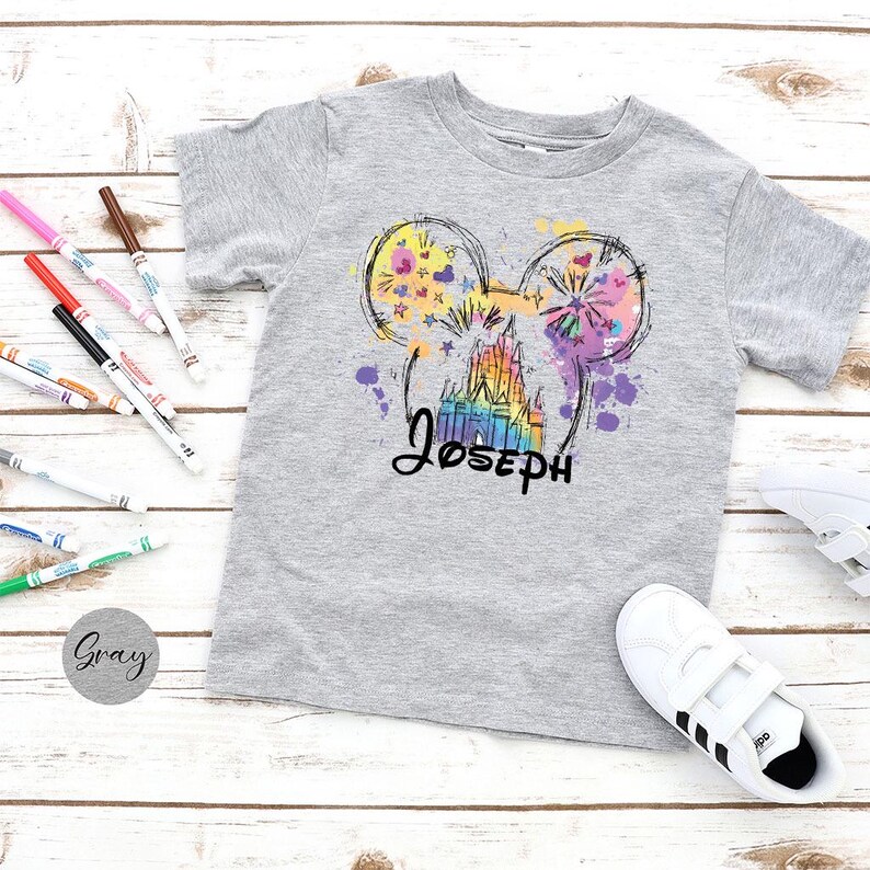 K&ouml;nnte beinhalten: Graues Kurzarm-T-Shirt mit einem farbenfrohen Aufdruck eines Schlosses und Mickey Mouse-Ohren mit dem Namen "Joseph" in schwarzer Schrift. Das Design enth&auml;lt Spritzer in Lila, Gelb und Rosa. Wei&szlig;e Turnschuhe und Marker sind ebenfalls sichtbar.