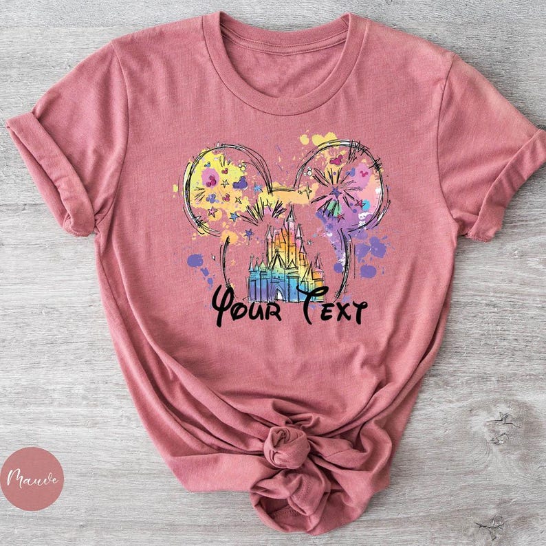 K&ouml;nnte beinhalten: Mauvefarbenes T-Shirt mit einer farbenfrohen Grafik eines Schlosses und Feuerwerks innerhalb von Mickey Mouse-Ohren. Der Text "Your Text" ist in einer Schrift unter dem Schloss geschrieben. Das Shirt ist unten geknotet.