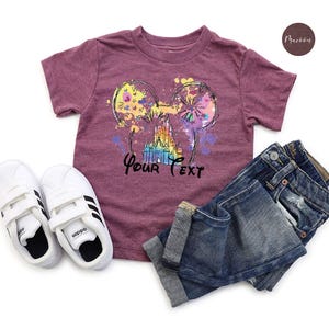 K&ouml;nnte beinhalten: Burgunderfarbenes T-Shirt mit einem farbenfrohen Aufdruck von Mickey Mouse Ohren, einem Schloss und dem Text "Your Text". Das Shirt wird mit blauen Jeans und wei&szlig;en Adidas-Sneakern kombiniert.