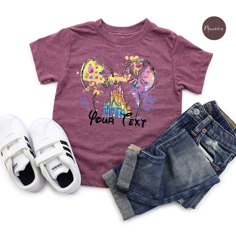 K&ouml;nnte beinhalten: Burgunderfarbenes T-Shirt mit einem farbenfrohen Aufdruck von Mickey Mouse Ohren, einem Schloss und dem Text "Your Text". Das Shirt wird mit blauen Jeans und wei&szlig;en Adidas-Sneakern kombiniert.