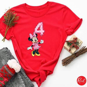 K&ouml;nnte beinhalten: Ein rotes T-Shirt mit einer Minnie Mouse-Grafik, die einen rosa Ballon mit der Zahl 4 h&auml;lt. Minnie Mouse tr&auml;gt ein rosa gepunktetes Kleid und eine Schleife. Das Wort "Red" befindet sich in einem Kreis unten rechts.