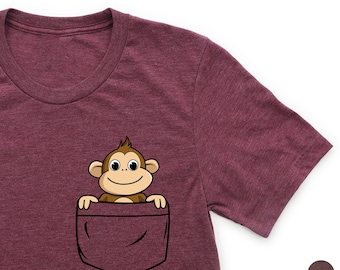 Puede incluir: Una camiseta de color granate con un bolsillo que muestra un mono de dibujos animados asomándose. El mono tiene pelo marrón, una cara sonriente y mira hacia la derecha.