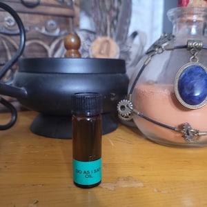 May include: A small brown glass bottle with a black cap and a teal label that reads "DO AS I SAY OIL." The bottle is on a wooden surface, with a black cauldron and a decorative glass jar with a blue pendant in the background.