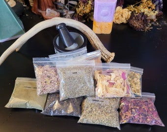 May include: A collection of dried herbs and flowers in clear plastic bags labeled with their names, including sage, rose petals, and dandelion leaf. The bags are arranged on a dark surface with a mortar and pestle and a purple and orange candle in the background.