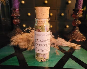 Puede incluir: Una pequeña botella de vidrio con tapón de corcho, llena de flores secas. La botella tiene una etiqueta que dice "Imaginarium Obscura" y "hydrangea". El fondo incluye una superficie verde, hierba seca y velas moradas.