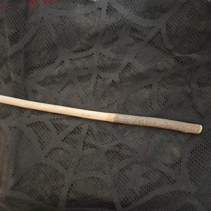 May include: A wooden wand featuring a smooth, light-colored tapered tip that transitions into a textured, dark brown bark-covered handle. The wand is positioned horizontally against a black fabric background with a spiderweb pattern.