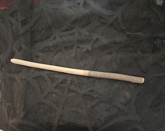 Könnte beinhalten: Ein Zauberstab aus Holz mit einer glatten, hellen und spitz zulaufenden Spitze, die in einen strukturierten, dunkelbraunen, rindenbedeckten Griff übergeht. Der Stab liegt horizontal auf einem schwarzen Stoff mit Spinnennetz-Muster.