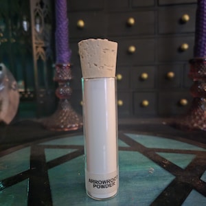 May include: A clear glass cylindrical vial filled with white arrowroot powder, sealed with a light brown cork stopper. A white label is affixed to the lower portion of the glass, featuring the black printed text ARROWROOT POWDER.