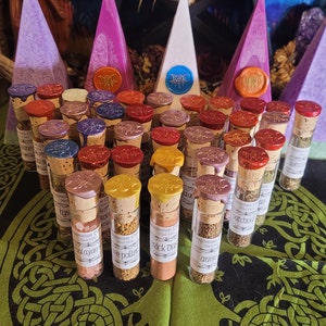 May include: A collection of small glass bottles with cork stoppers, filled with various herbs and spices. The bottles are arranged in rows on a green and brown patterned surface. The bottles are labeled with names such as "black oat", "sage pollen", and "anise".