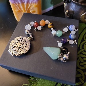 May include: A silver pendulum with a decorative mandala design and a green crystal point. The pendulum is attached to a chain with various coloured beads, including red, orange, yellow, green, blue, and purple.