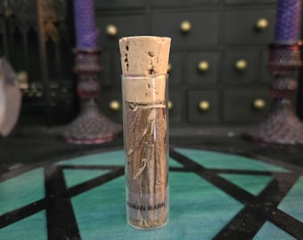 May include: A clear glass cylindrical vial filled with brown pieces of rowan bark. The vial is sealed with a light brown cork stopper and features a black label near the base with the text ROWAN BARK printed in clear, capitalized black letters.