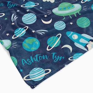 Può includere: Una coperta in pile blu a tema spaziale. La coperta presenta pianeti, stelle, un razzo, un UFO e il nome "Ashton Tyr".