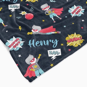 May include: A dark blue fleece blanket with a superhero theme. The blanket features a repeated pattern of a cartoon superhero boy wearing a red cape and a mask. The blanket also features stars, lightning bolts, and speech bubbles with the words "Super Boy", "Zap!" and "Boom!" The blanket is personalized with the name "Henry".