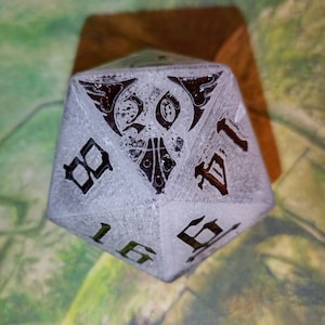 May include: A white, frosted, 20-sided die with black numbers and a detailed design on one side. The die is resting on a wooden surface.