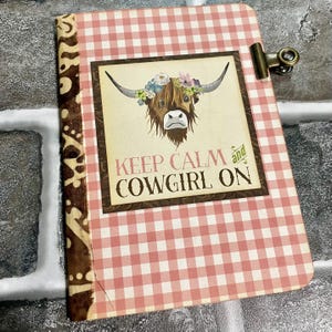 May include: A rectangular notebook with a pink and white gingham cover, a brown patterned spine, and a metal clip on the right edge. The center features a graphic of a highland cow wearing a floral crown with the text KEEP CALM and COWGIRL ON.