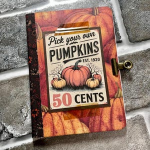 May include: A notebook with a pumpkin-themed cover featuring the text "Pick your own PUMPKINS" and "50 CENTS". The notebook has a decorative spine and a metal clip closure. The background is a collage of orange pumpkins.