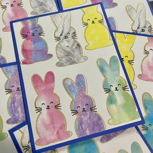 May include: Cards featuring watercolor bunny illustrations in various colors, including pink, purple, yellow, and blue. Each bunny has a gold outline and a smiling face with whiskers. The cards have a white background with a blue border.