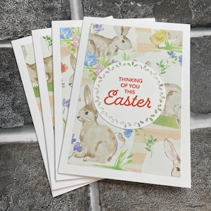 May include: Set of four blank greeting cards with a watercolor design of bunnies, flowers, and a pastel background. The cards have a white border and a circle with the text "Thinking of you this Easter" in red.