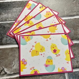 May include: A stack of rectangular cards with a pink border. The cards feature a repeating pattern of yellow chicks, flowers, and blue eggs on a white background. The cards are arranged in a fan shape.