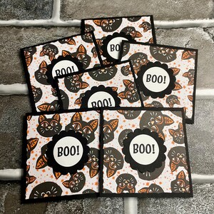 May include: A collection of Halloween-themed greeting cards. Each card features a black cat illustration on a patterned background with orange accents. The cards have a black scalloped circle with the word "BOO!" in white.