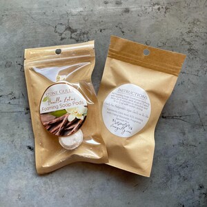 May include: Two brown paper pouches of Stone Gully Vanilla Lotus Foaming Soap Pods. One pouch has a label with a vanilla flower and pods. The other pouch has instructions. The pods are white and round.