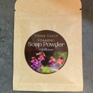 May include: A kraft paper packet of Stone Gully Foaming Soap Powder, Wildflower scent. The circular label features a floral image and text that reads "Makes 12oz". The packet is sealed at the top.