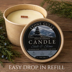 May include: A round, light brown paper container holding a cream-colored candle with a wooden wick. The lid features a black label with the text Stone Gully, Salt & Stone, and a mountain image. It includes the weight 5.1oz / 144g and text Easy Drop In Refill.