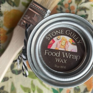 May include: A round metal container of Stone Gully Food Wrap Wax, 3oz/85g, with a brown label featuring an illustration of food wrapping. A paintbrush and a black and white twine are next to the container. The background has a floral pattern.