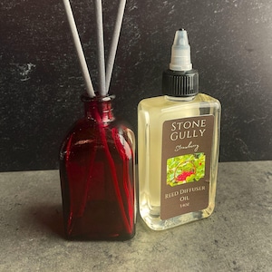 May include: A red glass reed diffuser bottle with gray reeds and a clear bottle of Stone Gully Strawberry Reed Diffuser Oil. The oil bottle has a brown label with a strawberry illustration and reads "14oz".