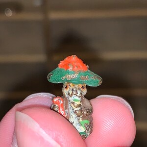 May include: A small, green and orange figurine of a person wearing a hat. The figurine is made of metal and has a detailed design.