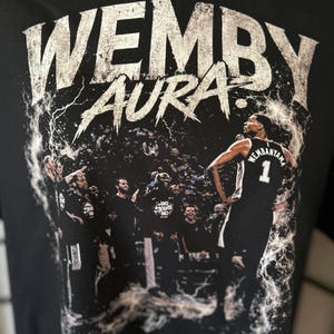 Könnte beinhalten: Schwarzes T-Shirt mit dem Text "WEMBY AURA" in weißer, abgenutzter Schrift. Das Design zeigt einen Basketballspieler in einem Trikot mit der Nummer 1 und dem Namen "WEMBANYAMA". Im Hintergrund ist eine Menschenmenge zu sehen.