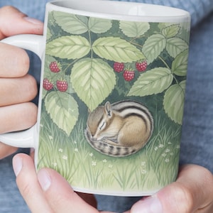 Op de afbeelding: Witte keramische mok met een ontwerp van een slapende chipmunk in het gras onder frambozenbladeren. Het ontwerp bevat rode frambozen, groene bladeren en witte bloemen. De mok heeft een wit handvat.