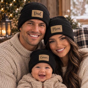 May include: Three black knit beanies featuring rectangular tan leather patches attached with gold-toned rivets. The patches are inscribed with the words Dad, Mom, and Connor in a dark cursive script font, centered on the front of each hat.