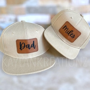 May include: Two beige baseball caps with brown leather patches. The patch on the left cap says "Dad" and the patch on the right cap says "Miles".