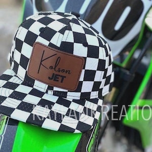 May include: A black and white checkered baseball cap with a brown leather patch that says "Kolson JET". The cap is sitting on a green and black dirt bike.