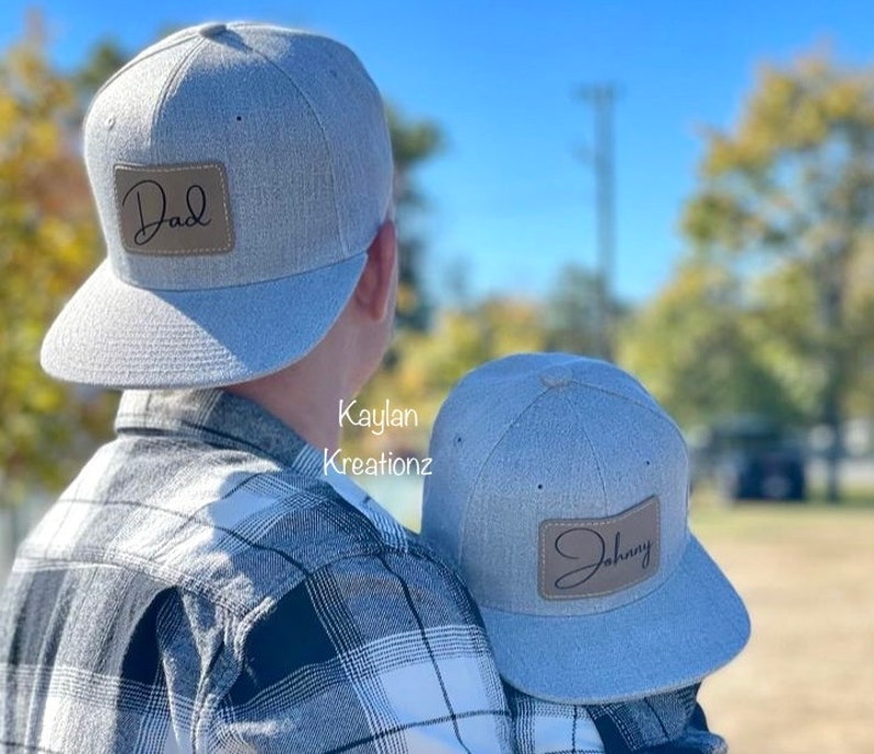 May include: Two gray baseball caps with brown leather patches. The larger cap has the word "Dad" written on the patch. The smaller cap has the word "Johnny" written on the patch.