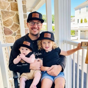 May include: A father and his two children are wearing black baseball caps with personalized leather patches. The father's cap says "Dad", the older child's cap says "Kava", and the younger child's cap says "Noah". The father is holding both children in his arms. The text "Kaylan Kreations" is visible in the image.